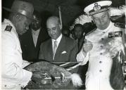 Fotografia de Américo Tomás, acompanhado por António Augusto Peixoto, em Quissico, recebendo presentes de um militar, por ocasião da visita de estado efetuada a Moçambique