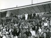 Fotografia de Américo Tomás, acompanhado por Gertrudes Rodrigues Tomás e por António Augusto Peixoto Correia, assistindo a uma parada desportiva no Estádio Santos Gil na cidade de Maputo, por ocasião da visita de estado efetuada a Moçambique